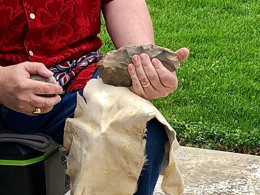 Archaeologist Nicholas Toth seated outdoors, striking a stone core with a hammerstone to make a replica handaxe.