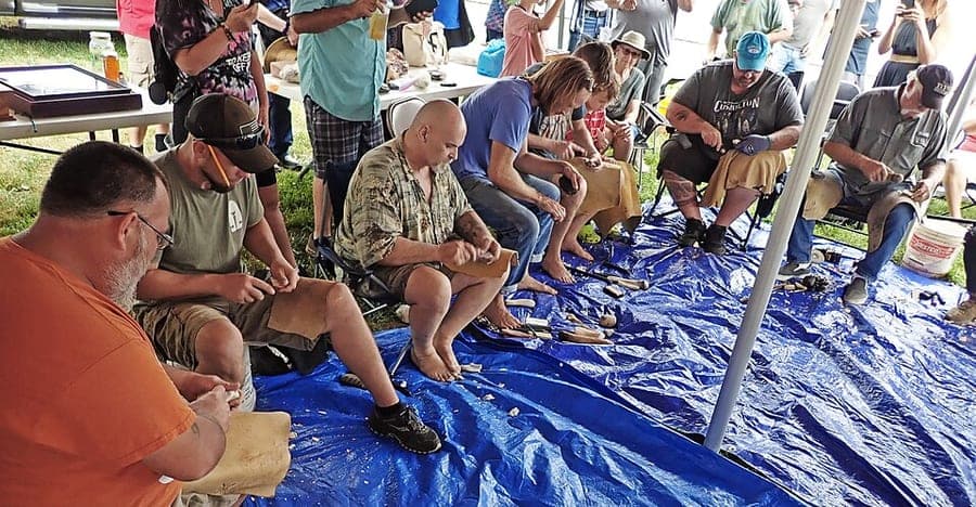 Several flintknappers seated at tables under a pavilion, working stone during a timed knapping competition at an Ohio flint festival.